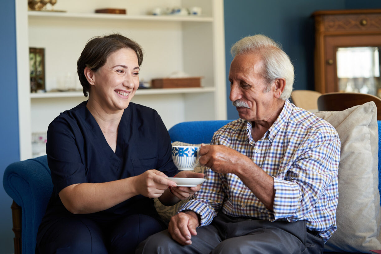 Professional helpful caregiver at nursing home. Health visitor and a senior man during home visit. Young caregiver in uniform and elderly man drinking tea