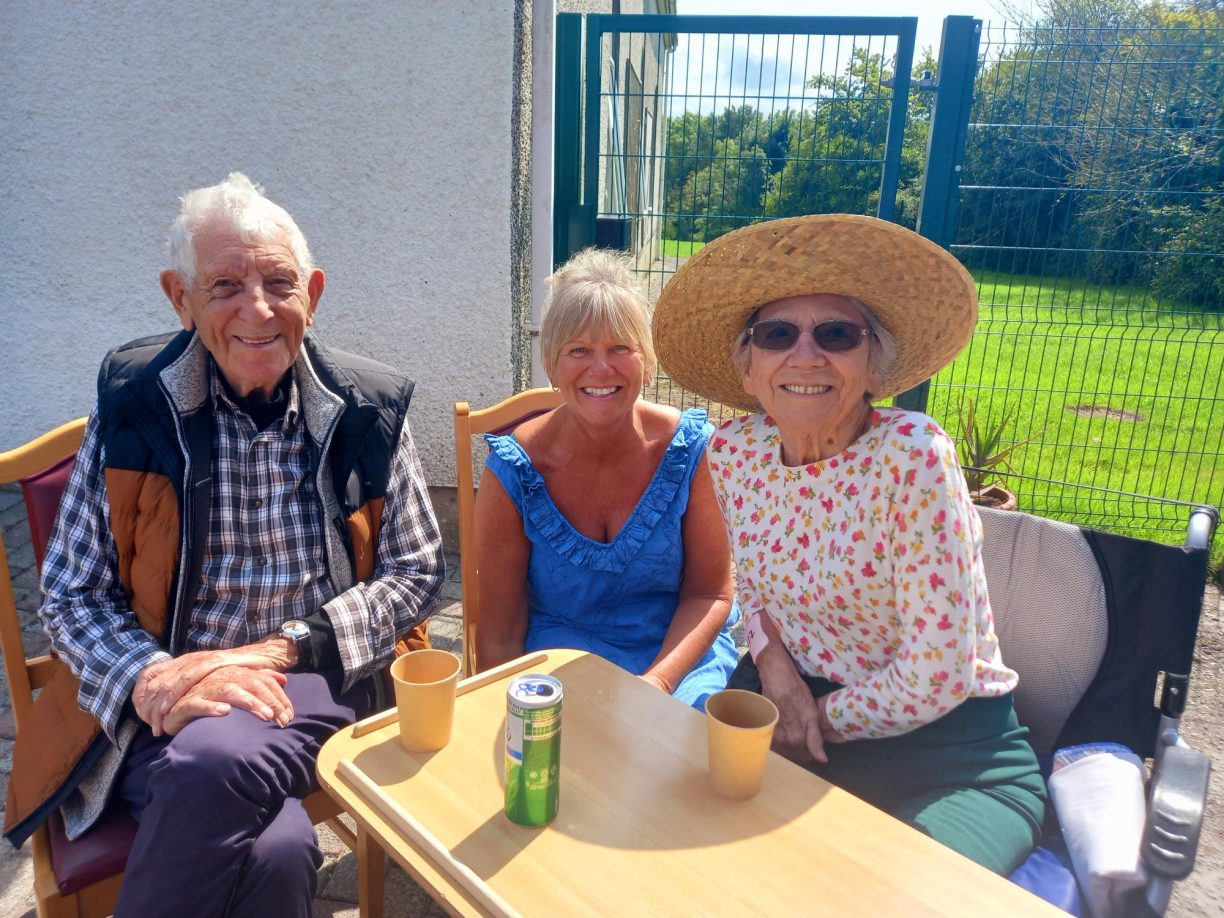Elderly female resident, Brenda, wearing a sunhat and sunglasses, sitting outdoors at a table with a man and a woman, who appear to be her family.