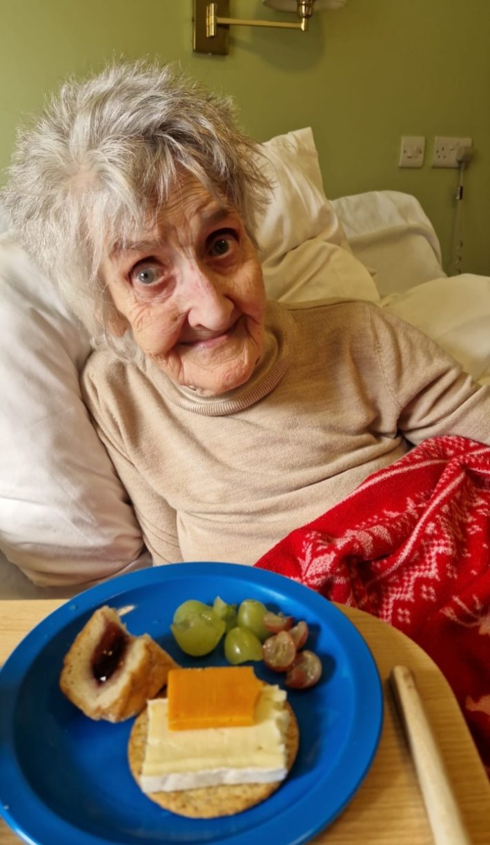 Elderly female resident Dodo sitting in bed and smiling, with a plate of cheese, crackers, and grapes in the foreground.