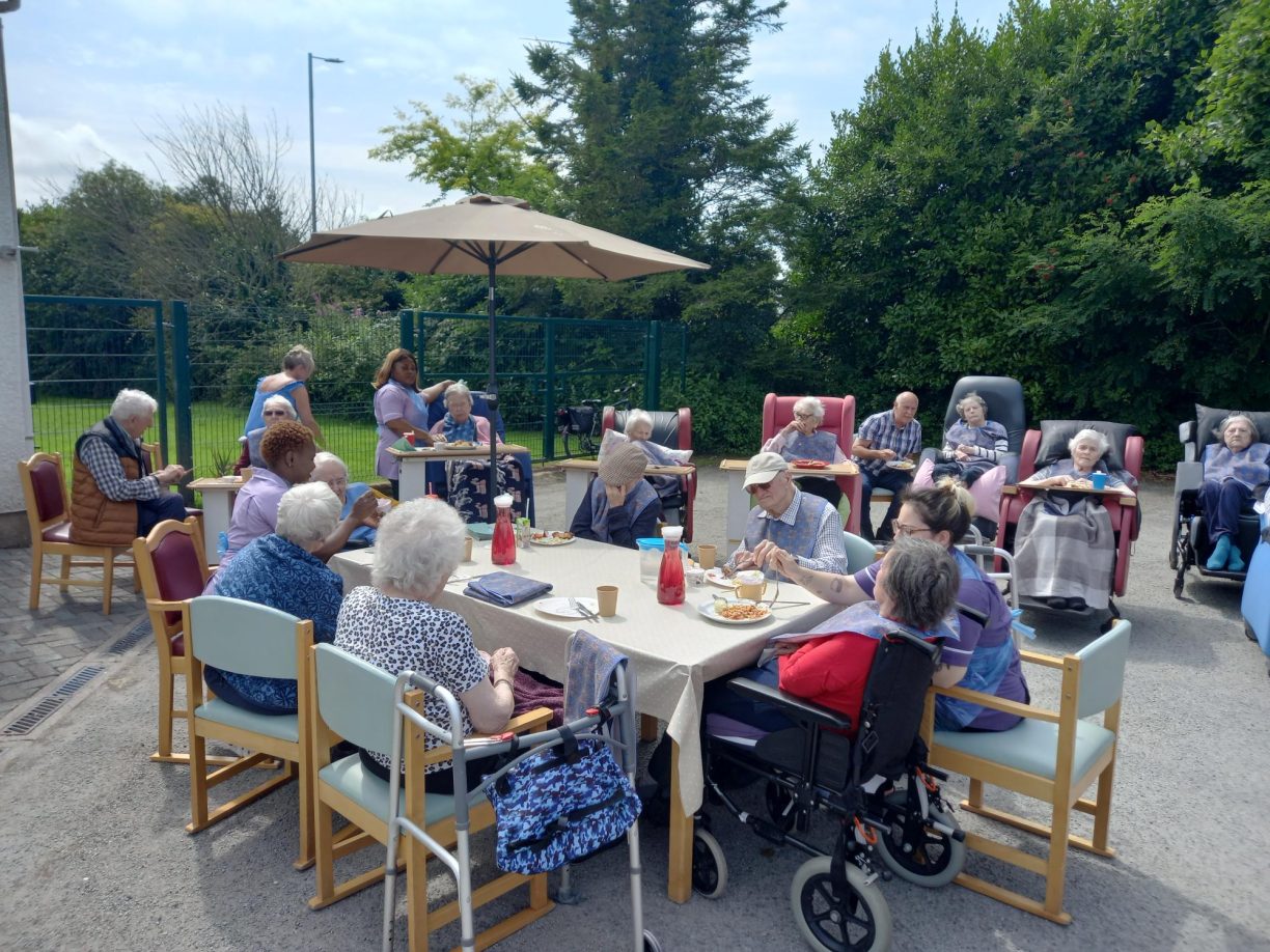 Numerous residents and care workers sitting outdoors enjoying a sunny afternoon meal or activity at Fairways Newydd.