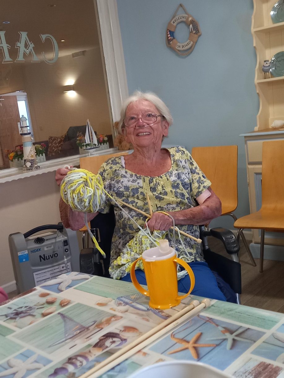 Elderly female resident, Brenda, sitting in a chair and smiling while holding a large ball of yellow and white yarn and knitting needles.