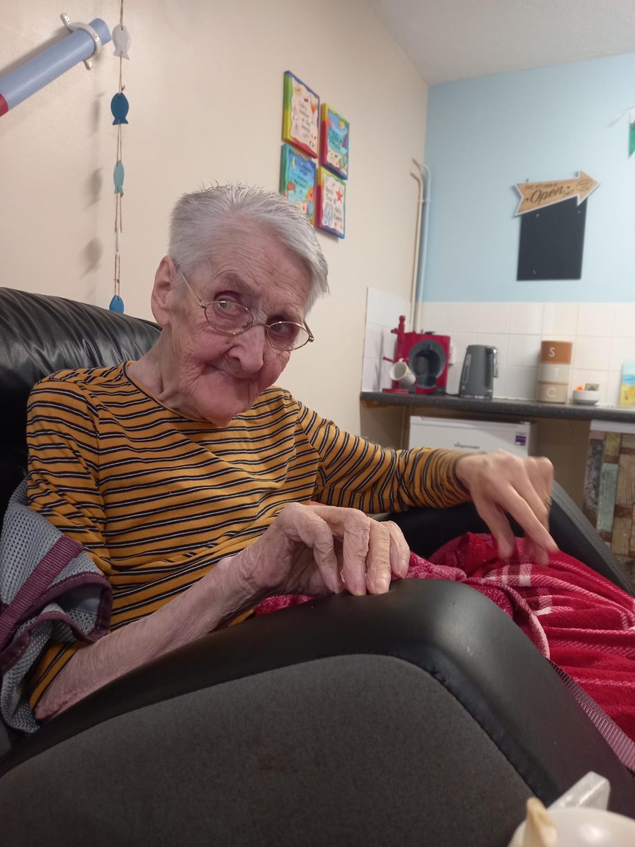 Elderly female resident, Dodo, sitting in a chair and smiling while holding a large ball of yellow and white yarn and knitting needles.