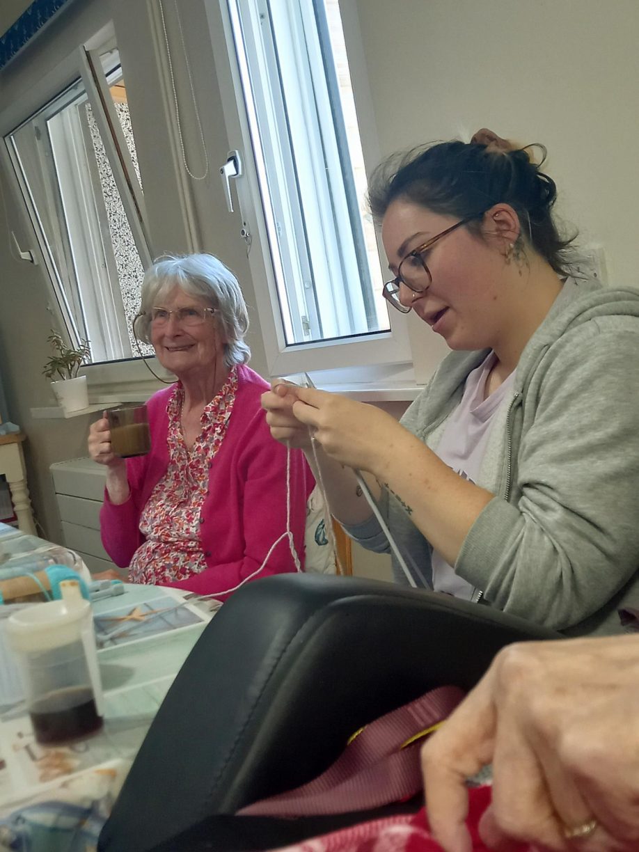 Care worker and elderly resident knitting together at a table, with the resident smiling and holding a mug.