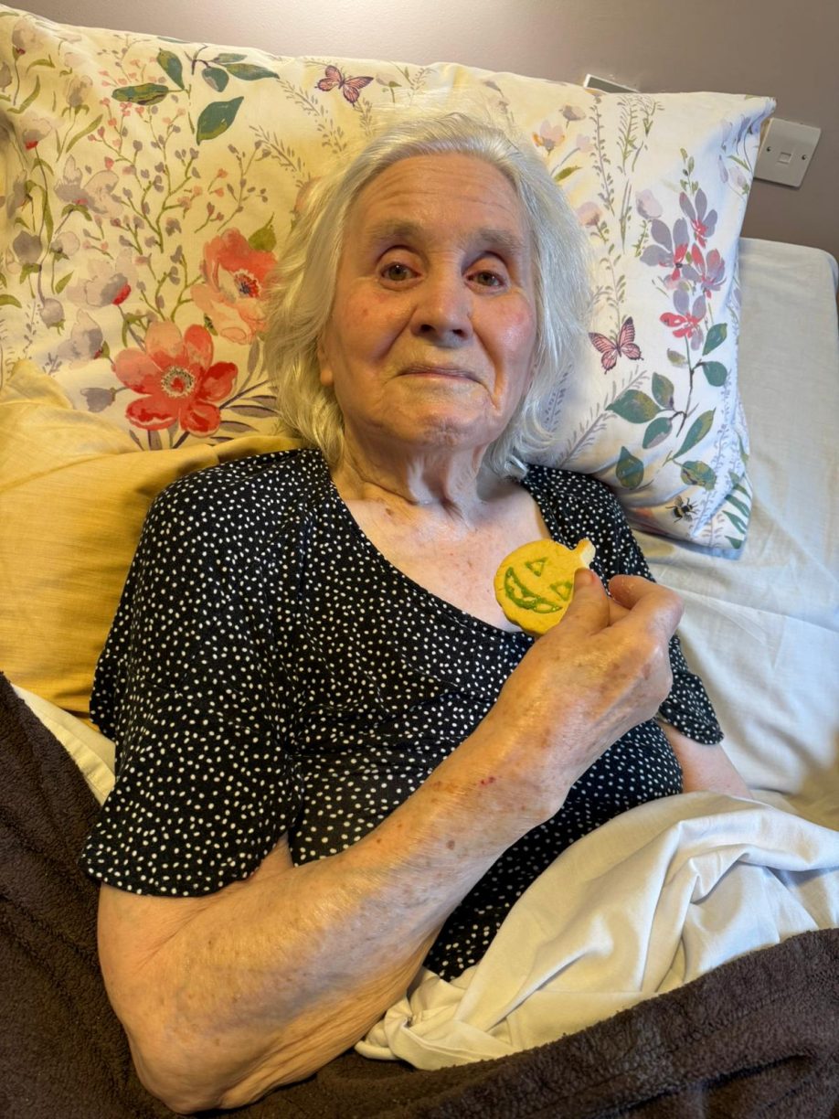 Elderly female resident resting in bed on a floral pillow, holding a jack-o'-lantern decorated Halloween cookie.