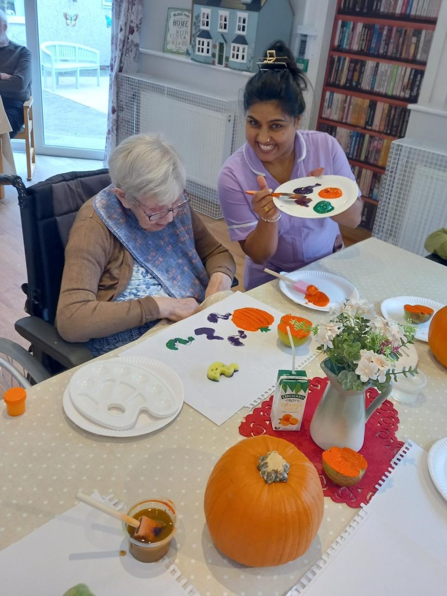 Care worker smiling while helping a resident in a wheelchair paint a pumpkin-shaped cookie and other crafts for Halloween.