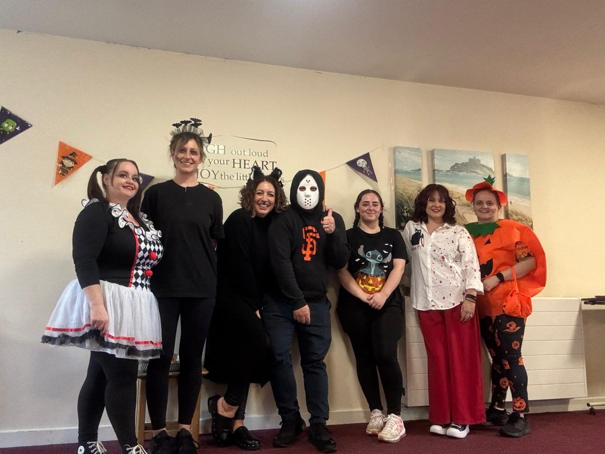 Seven care staff members dressed in assorted Halloween costumes, including a queen of hearts, a pumpkin, and Jason Voorhees.