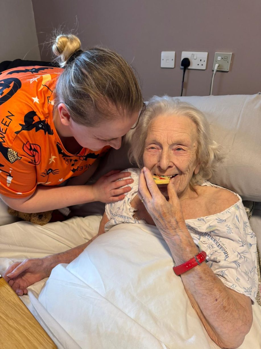 Care worker in a Halloween-themed shirt smiling down at a happy elderly resident eating a cookie in bed.