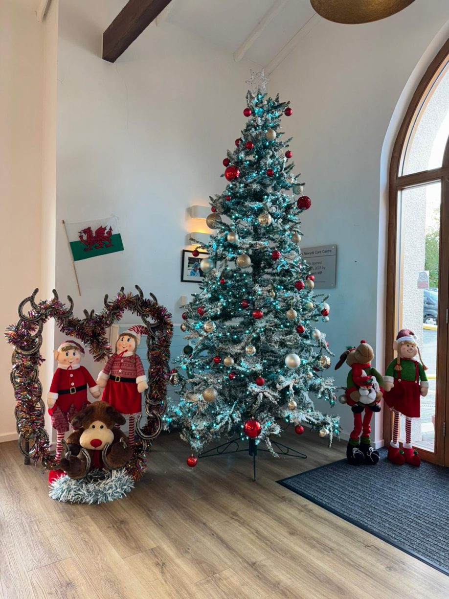 Tall, flocked Christmas tree decorated with red and gold ornaments next to decorative elf and reindeer plush figures.
